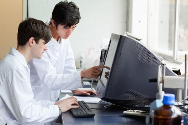 Two students collaborating on research in a university lab, surrounded by scientific equipment and monitors displaying data. One student is pointing at a graph on a screen, while the other is taking notes.
