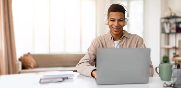 A young professional smiling while attending an online class on a laptop in a modern co-working space, with colleagues working in the background. The screen displays a video conference with a professor and other students.