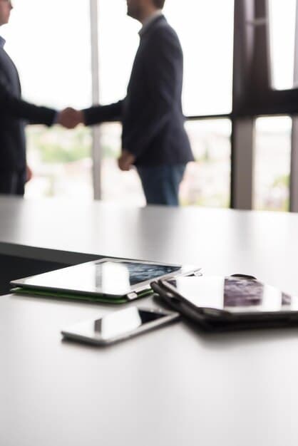 A close-up shot of two hands shaking firmly over a table with documents, symbolizing a successful business connection, with a blurred background of other professionals in a modern office setting.
