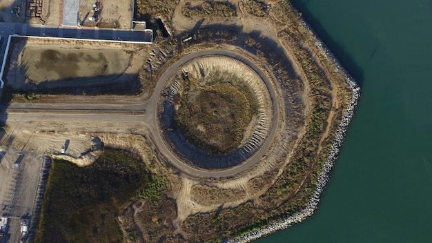 An aerial view of a large construction site for a hydroelectric dam. The site is bustling with heavy machinery, workers in hard hats, and concrete structures taking shape. The surrounding landscape is a mix of green forests and a winding river.