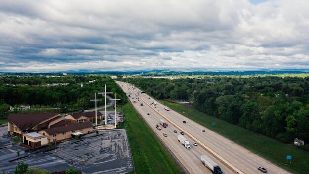 A panoramic view of a modern highway with several lanes, with cars and trucks moving in both directions, under a sunny sky. In the background, there are green hills and some buildings.